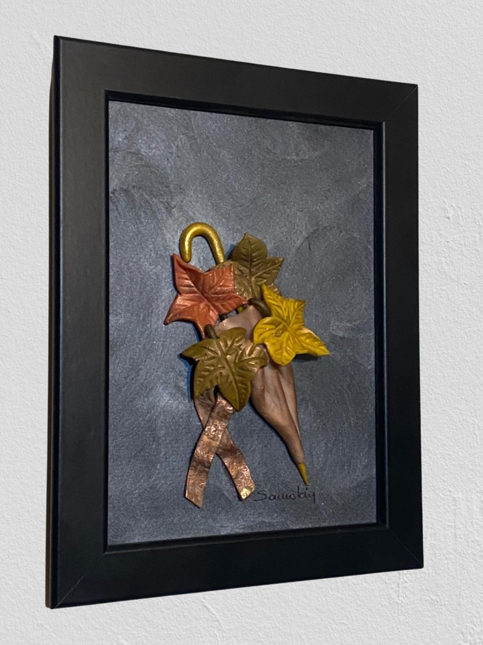 Autumn wall frame. It portraits four leaves in bronze, golden brown and olive green with an bronze umbrella.  All of the elements are in 3D form and crafted with polymer clay. The background is painted in industrial gray and the frame is black.