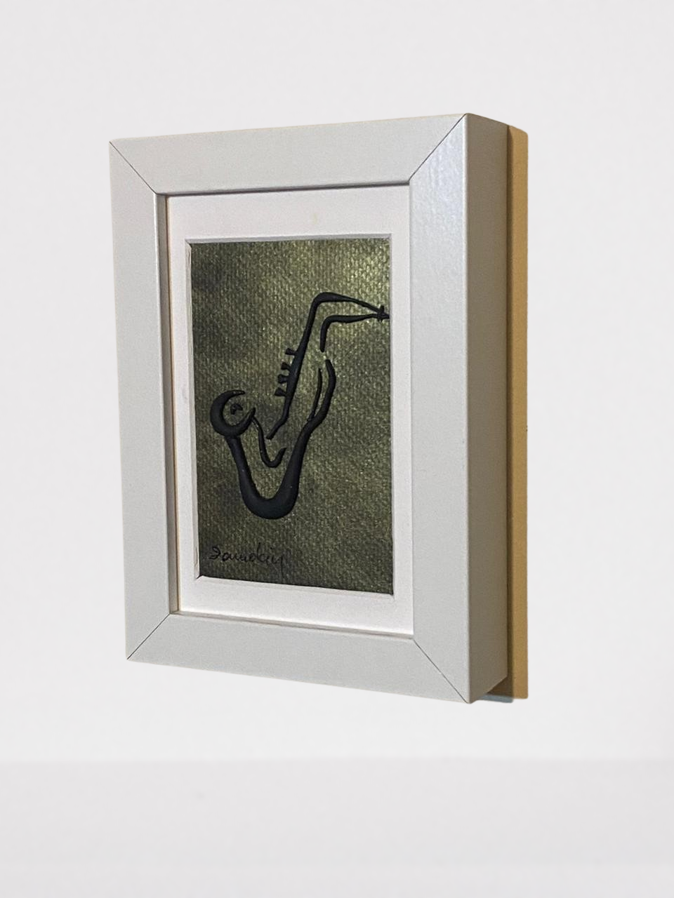 Handmade wall frame, featuring a 3D saxophone made with polymer clay on a golden painted canvas. The frame is white and made of wood.