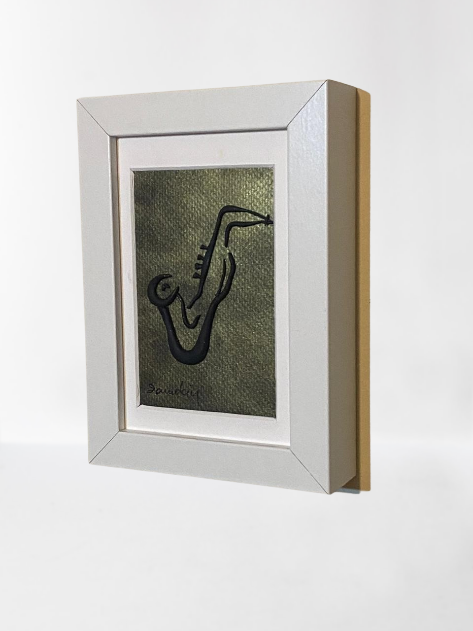 Handmade wall frame, featuring a 3D saxophone made with polymer clay on a golden painted canvas. The frame is white and made of wood.